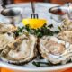 Delicious, fresh-shucked oysters in half shells sit on a chilled platter with a lemon garnish forked in the middle.