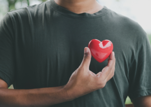 Close up of man in gray shirt holding a toy heart in front of his chest.