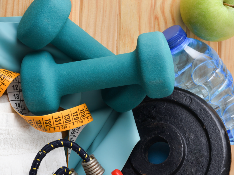 Close-up of fitness equipment on a light-colored wood surface to include blue hand weights, a black barbell weight, jump rope, bottled water, and a green apple.