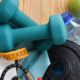 Close-up of fitness equipment on a light-colored wood surface to include blue hand weights, a black barbell weight, jump rope, bottled water, and a green apple.